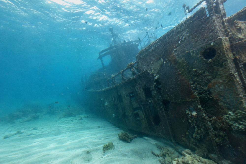 The dark hull of a sunken ship lying in clear blue water with fish nearby
