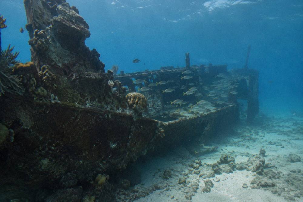A long sunken ship hull lying on its side on a sandy sea floor with fish swimming above it