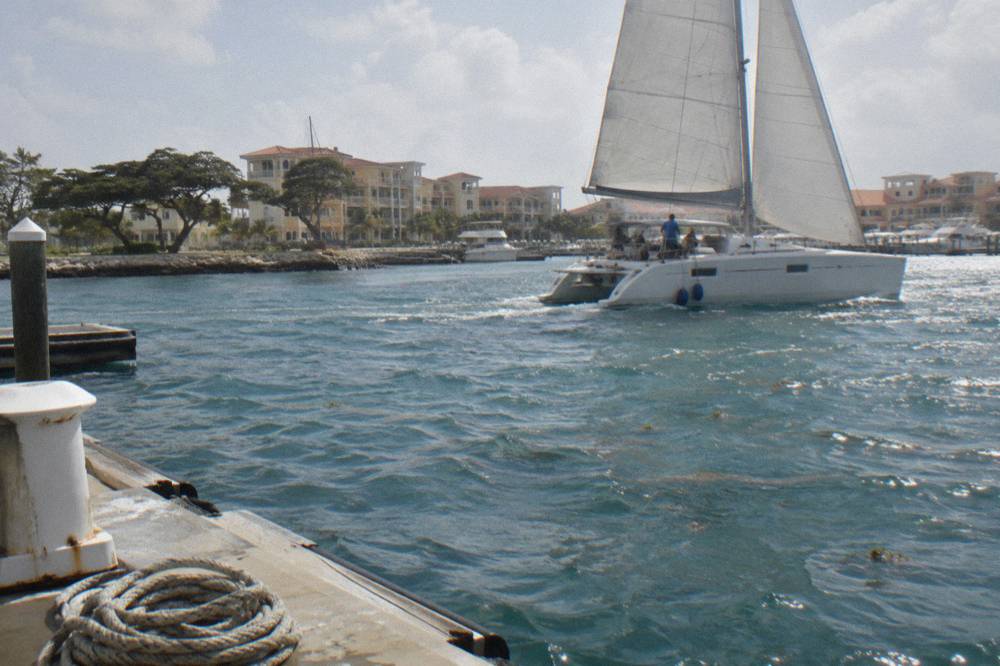 A catamaran with raised sails anchored near a reef in calm blue water