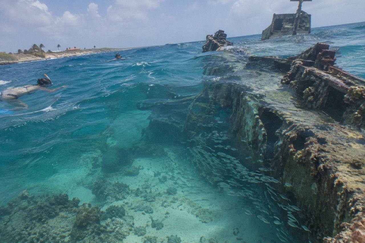 A sunken shipwreck lying on its side beneath calm blue Caribbean water near a coral reef
