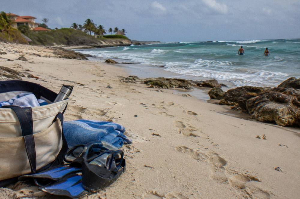 Beach bag with snorkel mask and fins on sand beside a folded towel under a clear sky