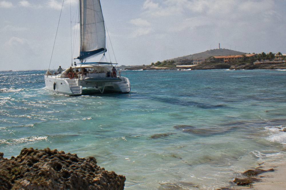A catamaran sailing along a calm coastline with clear shallow water over a sandy bottom