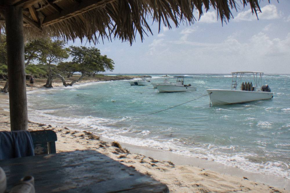 A sandy beach with calm shallow water and a line of white boats anchored offshore