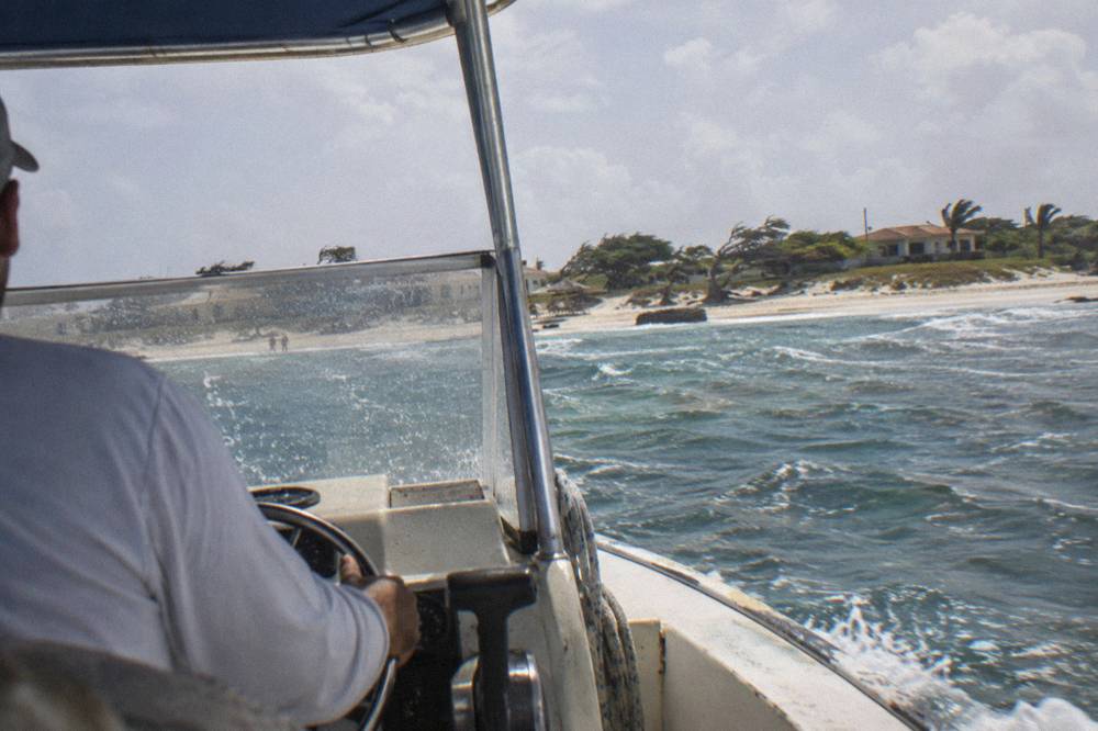 A captain at the wheel of a small boat cruising along a rocky coastline under a clear sky