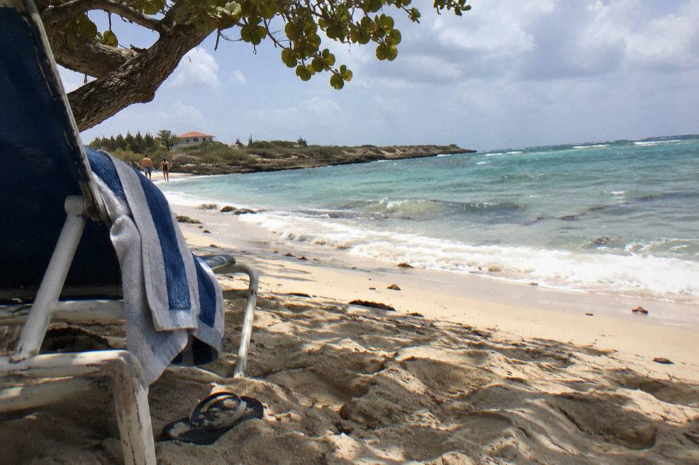 A shaded beach chair and towel on pale sand beside calm water