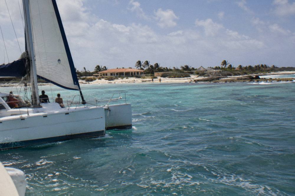 A catamaran under sail along a calm coastline with pale sand and clear water