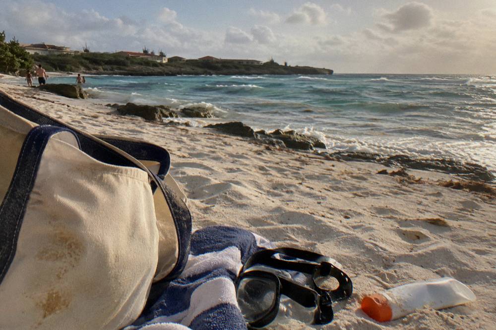 A beach bag, towel and snorkel mask arranged on sand beside calm water