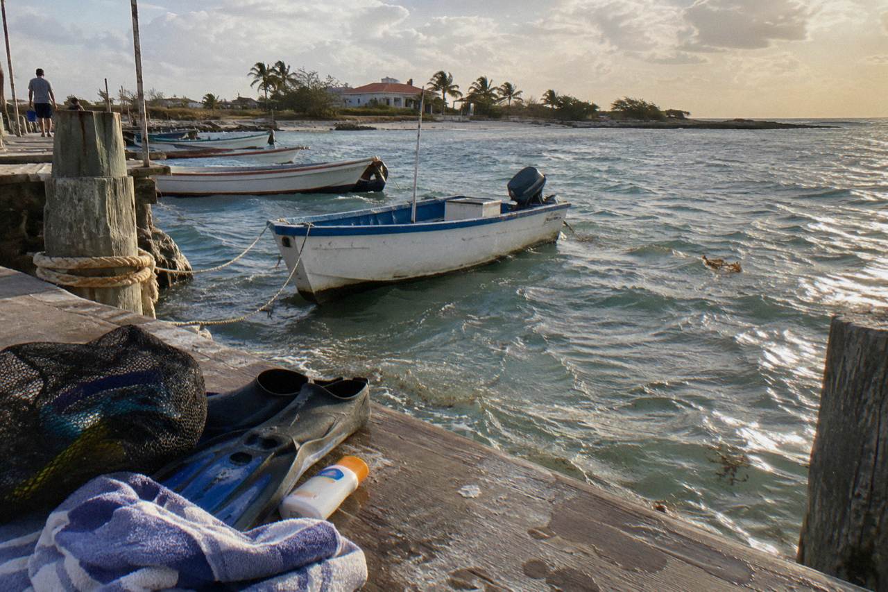 A mesh beach bag with snorkel mask, fins, towel, and sunscreen resting on a wooden dock beside calm turquoise water